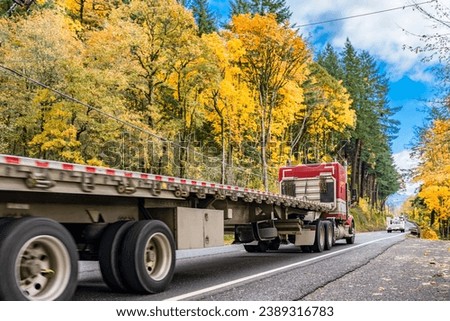 Red classic long hauler carrier big rig semi truck with extended cab transporting empty flat bed semi trailer running on the autumn narrow road with yellow trees on mountain hill in Columbia Gorge Royalty-Free Stock Photo #2389316783