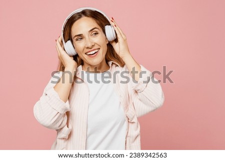 Young smiling happy woman she wear shirt white t-shirt casual clothes listen to music in headphones look aside on area isolated on plain pastel light pink background studio portrait. Lifestyle concept