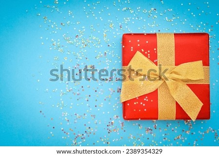 Blue christmas background with red gift box and bow. Textured cover of gift with gold ribbon and bow. Blue paper textured background has a lot of little stars. 