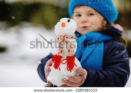 Cute little toddler girl making mini snowman and eating carrot nose. Adorable healthy happy child playing and having fun with snow, outdoors on cold day. Active leisure with children in winter.