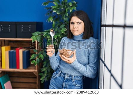 Young hispanic woman eating healthy whole grain cereals with spoon skeptic and nervous, frowning upset because of problem. negative person.  Royalty-Free Stock Photo #2389412063