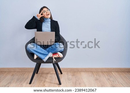 Young hispanic woman sitting on chair using computer laptop doing peace symbol with fingers over face, smiling cheerful showing victory 