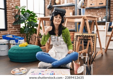 Hispanic woman painter sitting at art studio wearing virtual reality glasses smiling happy and positive, thumb up doing excellent and approval sign 