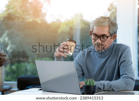 Asian senior man is holding cup of coffee while working his online business by using laptop, new edited.