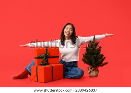Young Asian woman with gift boxes and Christmas tree on red background