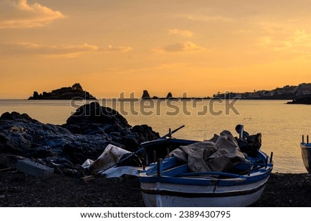 A traditional fishing beat is stranded on a pebble beach near Capo Mulini, in front of Isola Lachea, the famous Cyclops island in Homer's Odyssey, at sunset in Sicily, Italy Royalty-Free Stock Photo #2389430795