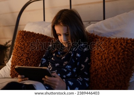 Cute little girl using tablet in bed viewing video. Young schoolgirl lying on comfortable pillows and reading ebook. Small lady browsing internet with her own pc at night. Child watching cartoon