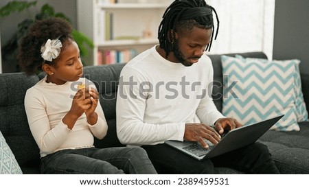 African american father and daughter sitting on sofa eating croissant using laptop at home