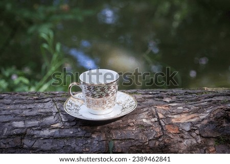 A cup of coffee with golden patterns, in a small saucer, stands on a log, against the backdrop of a picturesque river in the morning. Image for your design or creative illustrations.