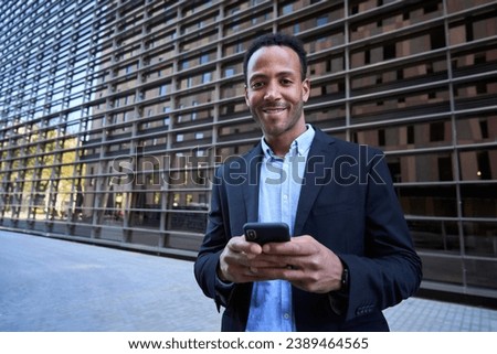 Portrait young African American professional man looking smiling at camera using mobile outside office center. Black business male in formal suit typing online message on phone on break from work.