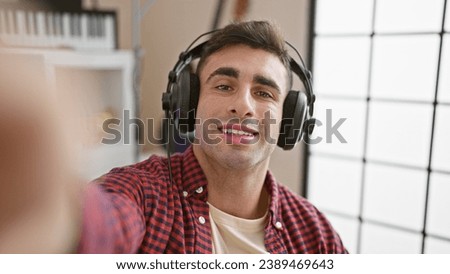 Confident young hispanic male musician, artist performing indoors, avidly listening to melody, passionately speaking on video call within music studio, headphones on, smiling while singing. Royalty-Free Stock Photo #2389469643