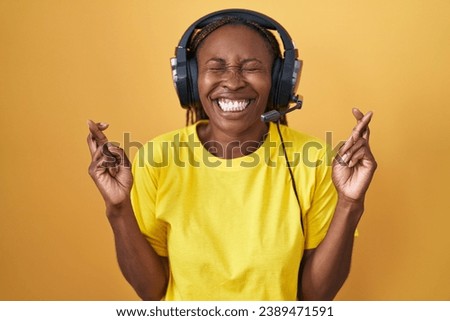 African american woman listening to music using headphones gesturing finger crossed smiling with hope and eyes closed. luck and superstitious concept. 