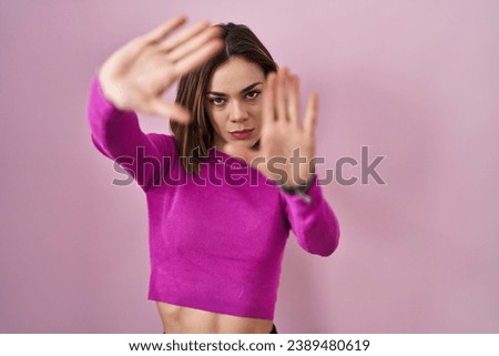 Hispanic woman standing over pink background doing frame using hands palms and fingers, camera perspective 