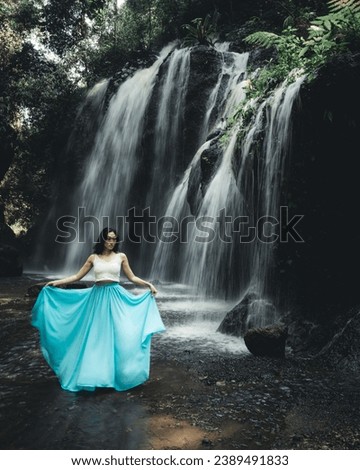 Asian woman posing near waterfall. Nature and environment. Motion water. Travel lifestyle. Charming woman wearing long blue skirt. Slim body. Copy space. Yeh Bulan waterfall, Bali. Motion photography.