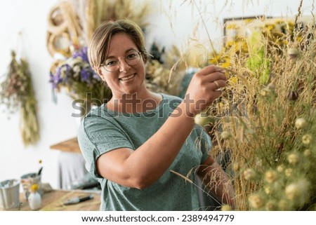 Smiling woman with glasses arranging dried plants in a floral workshop with hanging bouquets
