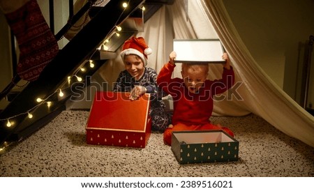 Two cheerful boys in pajamas playing in tepee tent under stairs and opening Christmas gifts and presents from Santa. Winter holidays, celebrations and party