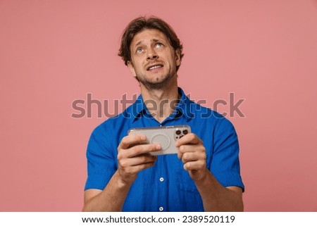 Emotional young man in blue shirt playing online game with smartphone while standing isolated over pink studio background