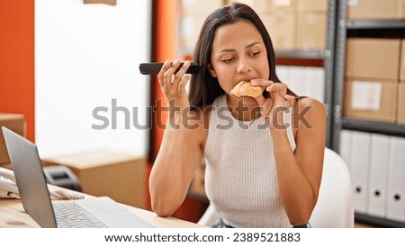 Young beautiful hispanic woman ecommerce business worker eating croissant working at office