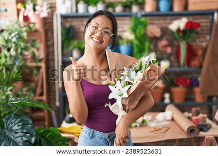 Asian young woman at florist shop holding bouquet of flowers smiling happy and positive, thumb up doing excellent and approval sign 