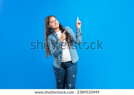 Advertisement concept, toothy smile. Happy Indian girl pointing fore fingers upwards for product advertisement on blue background