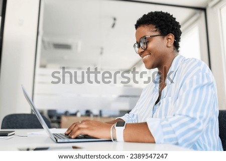 Optimistic african-american female office employee using laptop in modern coworking space, businesswoman in casual wear working on computer, watching at the screen and smiling, side view Royalty-Free Stock Photo #2389547247