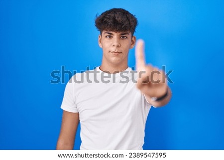 Hispanic teenager standing over blue background pointing with finger up and angry expression, showing no gesture 