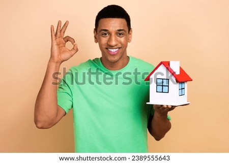 Photo portrait of attractive young man show okey sign hols little house dressed stylish green clothes isolated on beige color background
