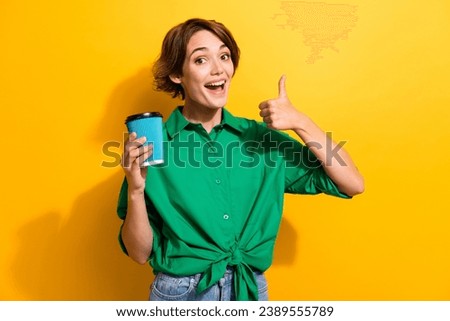 Photo of positive excited girl dressed green shirt rising enjoying coffee to go showing thumb up isolated yellow color background