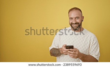 Cheerful young adult man in high spirits, touching his smartphone with a joyful smile, completely engrossed in a digitally communicated conversation, isolated against a refreshing yellow background