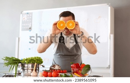 Chef Vlogger Showing Comic Orange Citrus Eye. Vegan Man with Cut Halves in Kitchen. Healthy and Fresh Juicy Fruit Ingredient. Fresh Tomatoes, Cabbage and Avocado for Lunch Looking at Camera Shoot