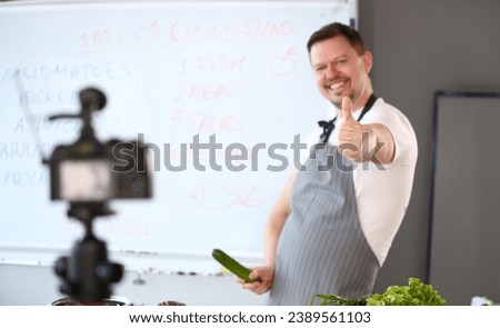 Cheerful Blogger Chef Showing Thumbup Gesture Joke. Cook in Apron Joking with Green Cucumber in Hand. Man Recording Culinary Healthy Recipe for Blog. Ripe Organic Cuke. Blackboard with Ingredient