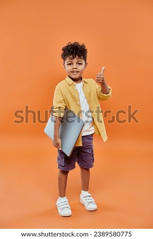 happy and curly african american boy showing like and holding laptop on orange background Royalty-Free Stock Photo #2389580775