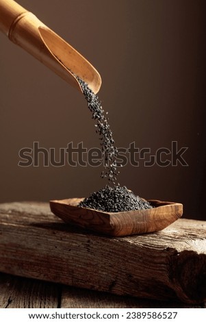 Grains of black sesame are poured with a wooden spoon in the wooden dish. Copy space. Royalty-Free Stock Photo #2389586527
