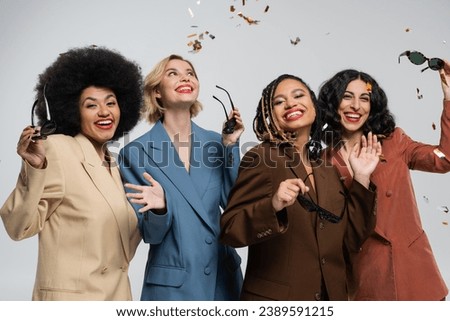 trendy multicultural girlfriends in colorful suits smiling under falling confetti, party time