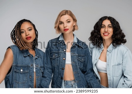 three attractive multiracial women in denim clothes looking at camera on grey, group portrait