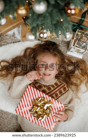 Adorable little girl of 5 years old with long curly hair and blue eyes opens gift boxes near the Christmas tree. Holidays. New Year. The winter vacation.