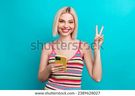 Photo portrait of pretty young girl show v-sign gold device nonverbal symbol wear trendy striped outfit isolated on cyan color background