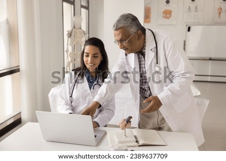 Two diverse doctors discussing online medical application, using laptop at workplace together. Indian elderly medical professional teacher, mentor training young Latin intern in clinic office Royalty-Free Stock Photo #2389637709