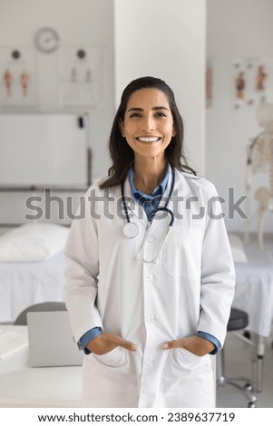 Happy beautiful young Hispanic doctor woman in white uniform coat standing in medical office, looking at camera with toothy smile. Cheerful medical professional enjoying occupation. Vertical shot Royalty-Free Stock Photo #2389637719