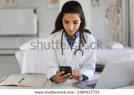 Serious young Latin practitioner using modern technology at workplace, typing on smartphone at table with laptop, keeping online connection with patients with medical healthcare application