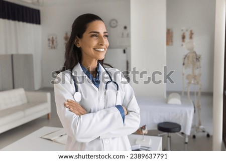 Happy young practitioner doctor posing in medical office, standing with hands crossed, looking away, smiling, thinking on successful therapy for patients, enjoying occupation Royalty-Free Stock Photo #2389637791