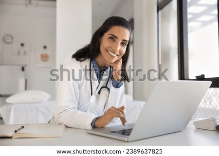 Cheerful attractive doctor woman professional portrait. Young Latin practitioner sitting at workplace table with laptop, looking at camera with toothy smile posing for shooting in surgery office Royalty-Free Stock Photo #2389637825
