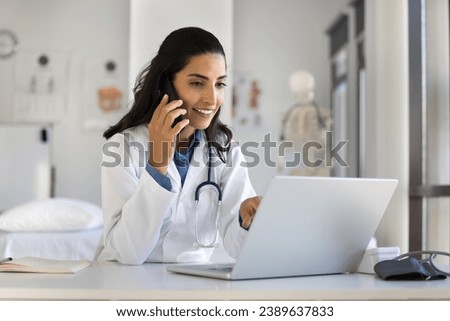 Young doctor woman talking to patient on mobile phone, smiling, using laptop at workplace, telling healthcare checkup test results, bringing good news, using modern technology for communication Royalty-Free Stock Photo #2389637833