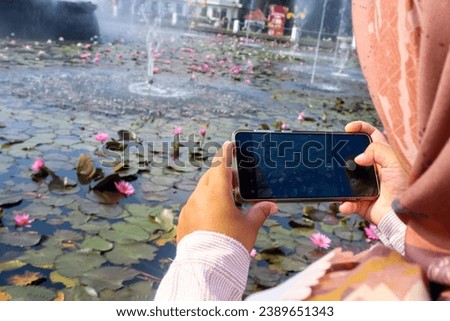 A woman with hijab take a picture using smartphone, blurred lotus flower in the background