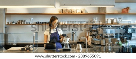 Portrait of cute girl student works as barista, holds POS credit card machine, standing at counter with terminal and brewing kit.