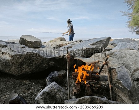 man fishing at the seaside while cooking fish on the wood campfire.
