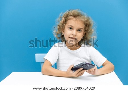 Cute little girl using smartphone and smiling while sitting at table on blue background. Happy girl holding phone in hands, online learning or homeschooling, listening to music or playing games.