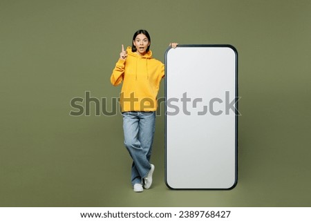 Full body young Latin woman she wears yellow hoody casual clothes big huge blank screen mobile cell phone smartphone with area point finger up isolated on plain pastel green background studio portrait