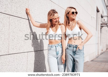 Two young beautiful smiling hipster female in trendy summer white top and jeans clothes. Carefree women posing in the street. Positive models having fun outdoors. Cheerful and happy. Near wall