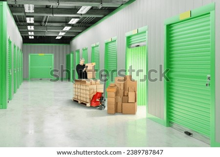 Man rents storage unit. Warehouse with gates to containers for safekeeping. Cardboard boxes near storage unit. Guy with phone in hallway of warehouse building. Storage unit inside building. Royalty-Free Stock Photo #2389787847